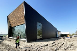 A woman in a visibility vest walks around a construction site with a wood clad building. 