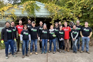 A group of people wearing matching T-shirts stand in front of a detached garage covered in vines. 