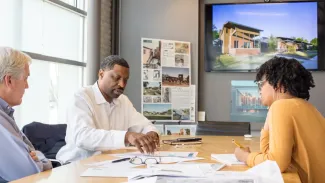 Three employees sit around a conference table with papers laid in front of them, on the screen behind them is an image of a building