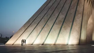 People in the shadow of a large sloped building made of parallel lines