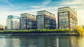 3D rendering of 4 rectangular buildings on a sunny day with a tree-lined walkway dividing it from a river 