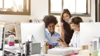 Three young employees smile as they all look over paperwork together at a desk in a sunny office