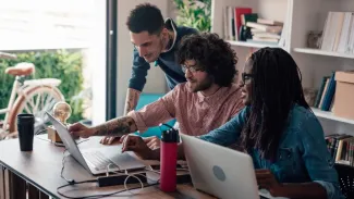 Three smiling employees sit and stand in front of a desk with two laptops open, behind them is a bookshelf