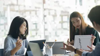 Three employees sit around the end of a conference table with laptops and paper, behind them is a brightly lit window