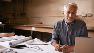 Older architect sitting at a desk with lots of plans and a laptop