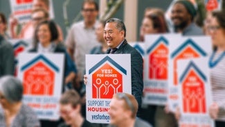 A group of people stand holding signs in favor of affordable housing. Douglas Ito stands center.