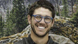 Headshot of a man with dark hair and glasses standing in front of a forest background