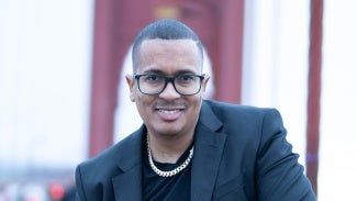 Headshot of a man with short hair in a black blazer wearing a chain necklace and glasses. A red bridge is visible in the background.