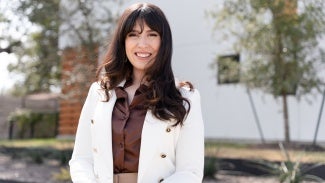 Headshot of a woman with long dark hair in a white blazer standing outside a building.