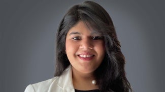 headshot of a woman with long dark hair and a white blazer in front of a grey background.