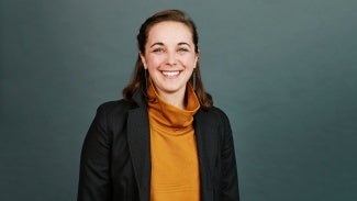 Headshot of a woman with long brown hair, wearing an orange sweater and a dark blazer.