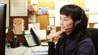 Woman with a headset on sitting at a desk looking at a computer monitor.