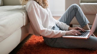 Woman siting on the floor against a couch with a laptop computer in her lap.