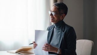 Women sits at table by a bright window, holding a pencil and a small stack of papers