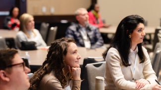 Participants sitting at table, listening intently to speaker. Two women in main focus, with other people blurred in the foreground and background.