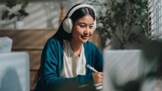 Lady sitting at a computer studying for exam, while taking notes and listening to headphones
