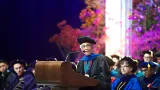 Woman in formal graduation attire speaking at a podium