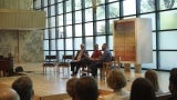 Three speakers sit on stage at a performance venue with a large Bank of windows behind them; a grand piano sits to the left