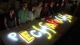A group of people stand behind a work table with a neon light sign on it.