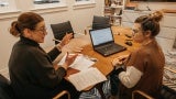 Two women wearing glasses sitting at a wooden office table and having a conversation. 