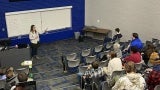 A woman with long hair and a white shirt giving a presentation in a lecture hall.