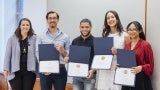 A group of five people standing in front of a grey wall. Four of them are holding diplomas.