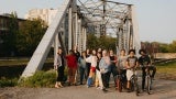 A group of people standing in front of a steel bridge. Three people on the right are standing with bicycles.