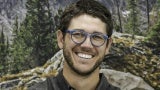 Headshot of a man with dark hair and glasses standing in front of a forest background