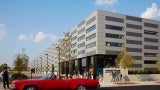 School exterior At day. The school has an alternating color facade of Gray and beige. There is a red car in the foreground. 