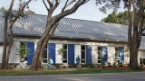 Building exterior with white cladding and blue shutters. There are three large trees in the foreground