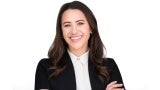 A woman with long dark hair in a black blazer stands in front of a white background.