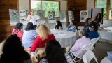 A woman gives a presentation to a large group of people sitting at round tables. 