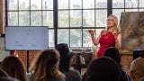 A woman gives a presentation to a large audience. There is a wall of windows behind her. 