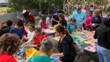 A large group of children work on art projects outside at various tables. There is a playground in the background.