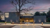 External shot of a grey community building from the parking lot at dusk. There is a bare tree in the foreground.