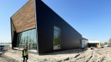 A woman in a visibility vest walks around a construction site with a wood clad building. 