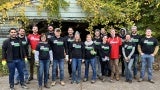 A group of people wearing matching T-shirts stand in front of a detached garage covered in vines. 