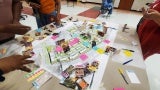 Photo of a table with an architectural plan on it. The plan is covered in sticky notes. You can see the hands of four people standing around the table out of frame. 