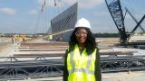 A woman in a visibility vest and a hard hat stands on a construction site. 