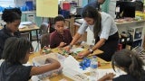 Four children sit around a classroom table with an instructor in the center. The instructor is helping them with an art project.