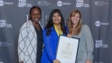 Three women in front of a step and repeat backdrop at an event. The woman in the center is wearing a medal and holding a framed certificate.