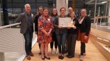 A group of people posed for a photo in an atrium. The person at the center of the group is holding a certificate. 