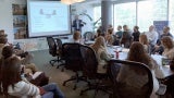 View of a classroom from the back. There is a boardroom table in the middle of the room with rolling chairs. Students sit at the table as well as along the sides of the room. A teacher is standing in the front of the classroom next to a projection screen giving a lecture.