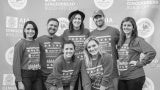 A group of seven individuals in matching t-shirts pose in front of an event step and repeat.