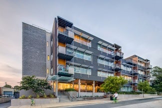 External view of a grey brick apartment building from the street at dusk. There are trees along the sidewalk.