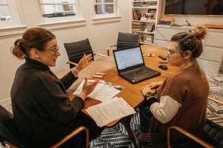 Two women wearing glasses sitting at a wooden office table and having a conversation. 