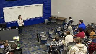 A woman with long hair and a white shirt giving a presentation in a lecture hall.