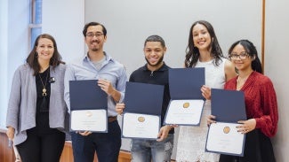 A group of five people standing in front of a grey wall. Four of them are holding diplomas.