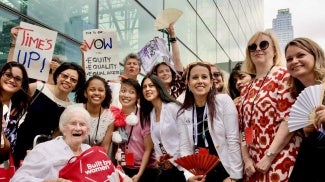 A group of diverse women stand in front of a glass building holding wage equality signs and paper fans.