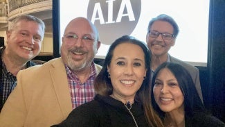 A group of five individuals take a selfie in front of a projection screen at an event.